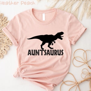Op de afbeelding: Een roze t-shirt met een zwart dinosaurus silhouet en de tekst "AUNTSAURUS" in zwart.