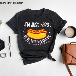 May include: Dark grey heather t-shirt with a hot dog graphic and the text "I'm just here for the weiners". The graphic includes red, white, and blue stars and a string of American flags.