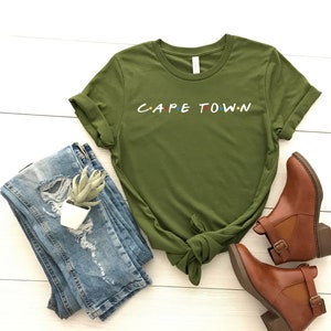 May include: A green t-shirt with the words "CAPE TOWN" in white letters with colorful dots. The shirt is folded and tied in a knot. The shirt is laying on a white wooden surface with blue jeans and brown boots.