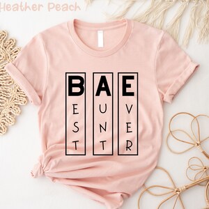 Op de afbeelding: Een roze T-shirt met het woord "BAE" in zwarte letters. De letters zijn gemaakt van kleinere letters die "Best, Unforgettable, Ever" spellen.