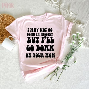 May include: Light pink t-shirt with the text "I MAY NOT GO DOWN IN HISTORY BUT I'LL GO DOWN ON YOUR MOM" in black, retro-style font. The shirt is folded, with a white mug and decorative elements in the background.