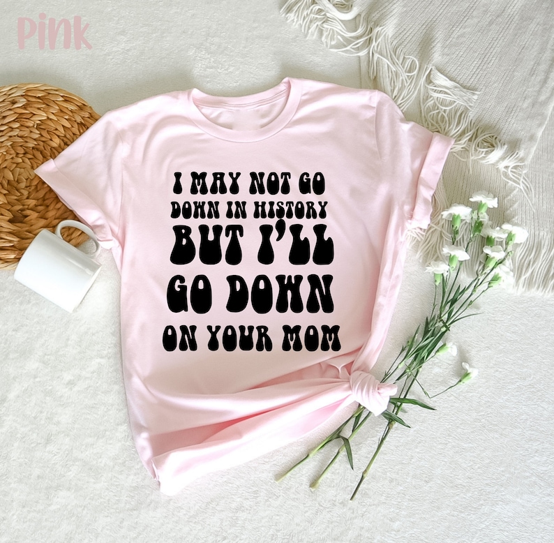 May include: Light pink t-shirt with the text "I MAY NOT GO DOWN IN HISTORY BUT I'LL GO DOWN ON YOUR MOM" in black, retro-style font. The shirt is folded, with a white mug and decorative elements in the background.