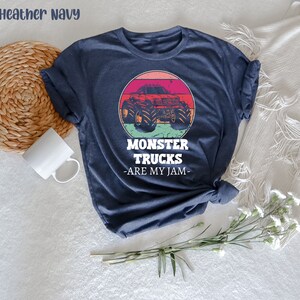 May include: Heather navy blue t-shirt featuring a vintage-style graphic of a monster truck in front of a sunset. The text "Monster Trucks - Are My Jam -" is printed below the graphic.
