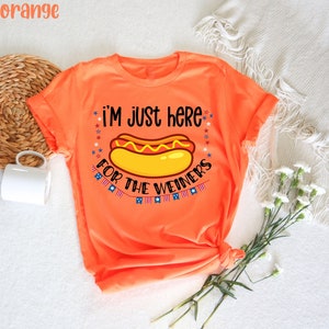 May include: Orange t-shirt with a hot dog graphic and the text "i'm just here for the weiners" with stars and American flags.