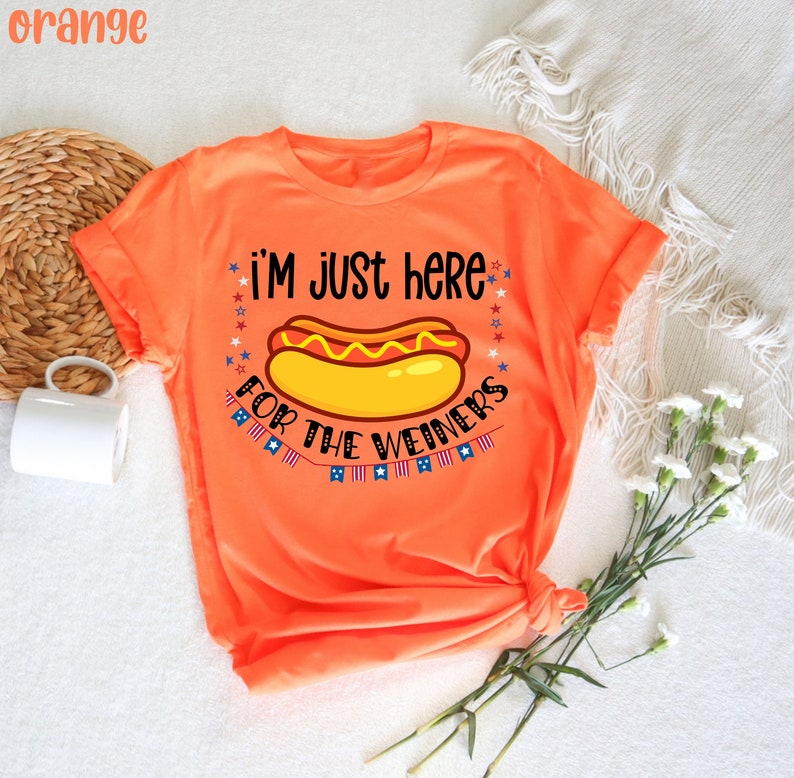 May include: Orange t-shirt with a hot dog graphic and the text "i'm just here for the weiners" with stars and American flags.