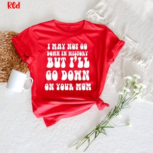 May include: A red t-shirt with the text "I MAY NOT GO DOWN IN HISTORY BUT I'LL GO DOWN ON YOUR MOM" in white, bold, retro-style font. The shirt is displayed on a white surface with a white mug and flowers.