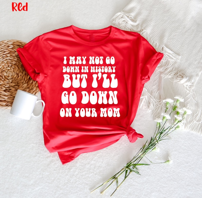 May include: A red t-shirt with the text "I MAY NOT GO DOWN IN HISTORY BUT I'LL GO DOWN ON YOUR MOM" in white, bold, retro-style font. The shirt is displayed on a white surface with a white mug and flowers.