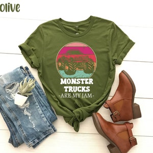 May include: Olive green t-shirt featuring a retro-style graphic of a monster truck in front of a sunset. The text "Monster Trucks - Are My Jam -" is printed below the graphic.
