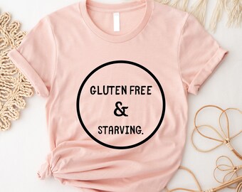 Könnte beinhalten: Ein rosa T-Shirt mit einem schwarzen Kreis-Motiv, auf dem "Gluten Free & Starving." steht.