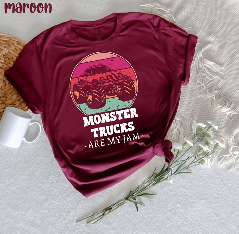 May include: A maroon t-shirt featuring a vintage-style graphic of a monster truck against a sunset backdrop. The text "Monster Trucks -Are My Jam- " is printed beneath the graphic.