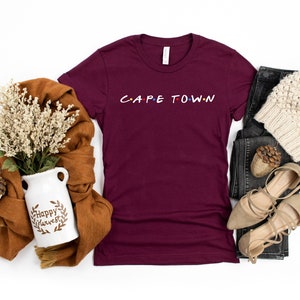 May include: A maroon t-shirt with the text "CAPE TOWN" in a stylized font. The shirt is laid flat on a white background with a brown scarf, a white ceramic vase with the text "Happy Harvest", a pair of brown sandals, a pair of black jeans, and a white knitted beanie.