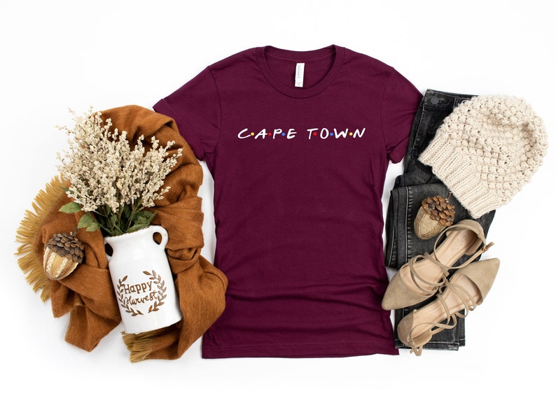 May include: A maroon t-shirt with the text "CAPE TOWN" in a stylized font. The shirt is laid flat on a white background with a brown scarf, a white ceramic vase with the text "Happy Harvest", a pair of brown sandals, a pair of black jeans, and a white knitted beanie.