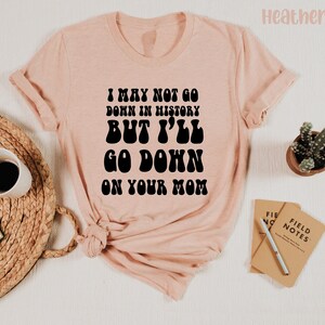 May include: Heather peach-colored t-shirt with the text "I MAY NOT GO DOWN IN HISTORY BUT I'LL GO DOWN ON YOUR MOM" in black. The shirt is styled with rolled-up sleeves and is laid on a woven mat with a coffee cup and a book.