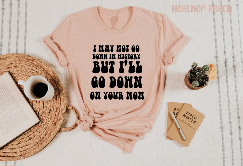 May include: Heather peach-colored t-shirt with the text "I MAY NOT GO DOWN IN HISTORY BUT I'LL GO DOWN ON YOUR MOM" in black. The shirt is styled with rolled-up sleeves and is laid on a woven mat with a coffee cup and a book.