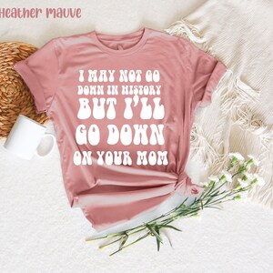 May include: Heather mauve t-shirt with the text "I MAY NOT GO DOWN IN HISTORY BUT I'LL GO DOWN ON YOUR MOM" in white. The shirt is knotted at the bottom and is displayed on a white surface with a mug and flowers.