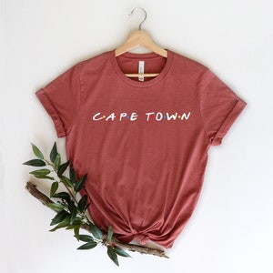 May include: A rust-colored t-shirt with the words "CAPE TOWN" in a stylized font with colorful dots.
