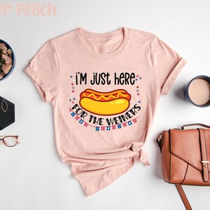 May include: A pink t-shirt with the text "I'm just here for the weiners" and a cartoon hot dog with mustard and ketchup. The text is surrounded by red, white, and blue stars and flags.