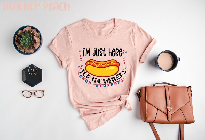 May include: A pink t-shirt with the text "I'm just here for the weiners" and a cartoon hot dog with mustard and ketchup. The text is surrounded by red, white, and blue stars and flags.