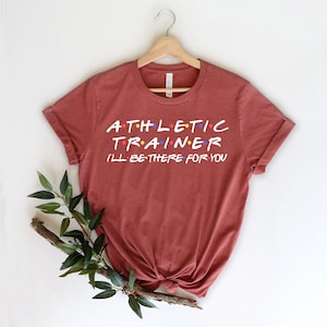 Pode incluir: Uma camiseta marrom-avermelhada com o texto "ATHLETIC TRAINER I'LL BE THERE FOR YOU" em branco com pontos coloridos. O texto é estilizado para se parecer com o logotipo da série Friends.