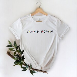 May include: White t-shirt with a black graphic that says "CAPE TOWN" in the style of the "Friends" logo. The graphic has colorful dots.
