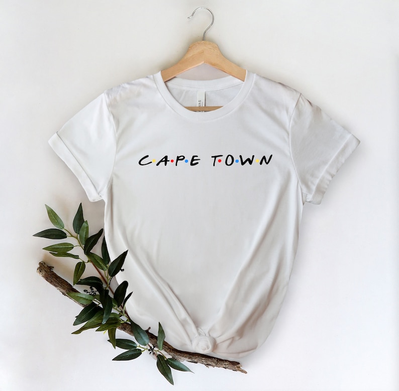 May include: White t-shirt with a black graphic that says "CAPE TOWN" in the style of the "Friends" logo. The graphic has colorful dots.
