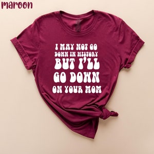 May include: Maroon t-shirt with white text that reads "I MAY NOT GO DOWN IN HISTORY BUT I'LL GO DOWN ON YOUR MOM". The shirt is folded, showing the rolled-up sleeves.