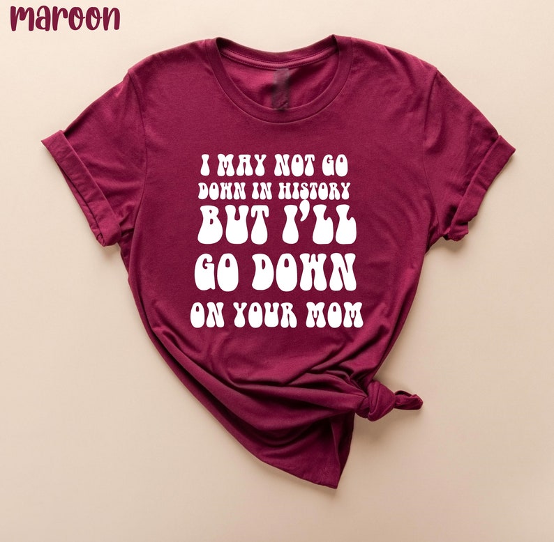 May include: Maroon t-shirt with white text that reads "I MAY NOT GO DOWN IN HISTORY BUT I'LL GO DOWN ON YOUR MOM". The shirt is folded, showing the rolled-up sleeves.