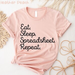 Könnte beinhalten: Ein rosa T-Shirt mit schwarzem Text, der "Eat. Sleep. Spreadsheet. Repeat." lautet.