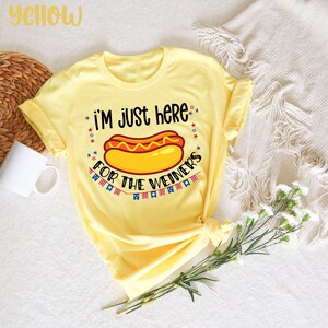 May include: Yellow t-shirt with a hot dog graphic and the text "I'm just here for the weiners". The graphic includes red, white, and blue stars and a string of flags.