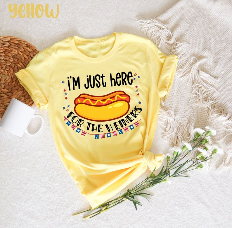 May include: Yellow t-shirt with a hot dog graphic and the text "I'm just here for the weiners". The graphic includes red, white, and blue stars and a string of flags.