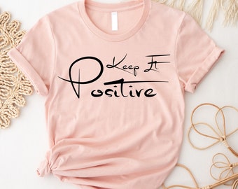 May include: A pink t-shirt with the text "Keep It Positive" in black lettering. The text is written in a stylized font with a circle around the "P" and a line through the "I".