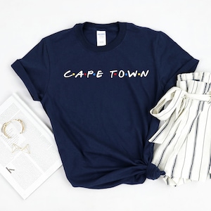 May include: A navy blue t-shirt with the text "CAPE TOWN" in white letters with colorful dots. The shirt is folded and laid on a white surface with a white and black striped skirt.