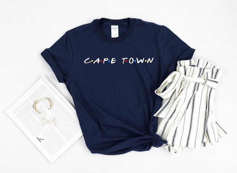 May include: A navy blue t-shirt with the text "CAPE TOWN" in white letters with colorful dots. The shirt is folded and laid on a white surface with a white and black striped skirt.