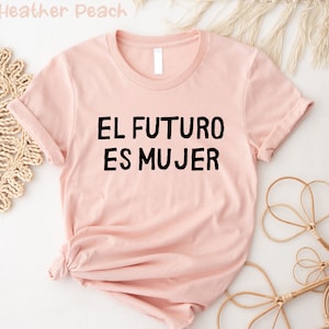 以下が含まれることがあります： 黒い文字で「EL FUTURO ES MUJER」とプリントされたピンクのTシャツです。