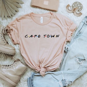 May include: A light pink t-shirt with the text "CAPE TOWN" in a stylized font with colorful dots. The shirt is laid out on a white fuzzy surface with a pair of light brown combat boots, a pair of light blue jeans, and a small brown clutch.