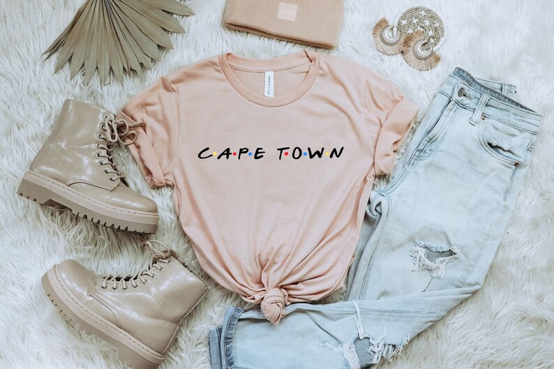 May include: A light pink t-shirt with the text "CAPE TOWN" in a stylized font with colorful dots. The shirt is laid out on a white fuzzy surface with a pair of light brown combat boots, a pair of light blue jeans, and a small brown clutch.