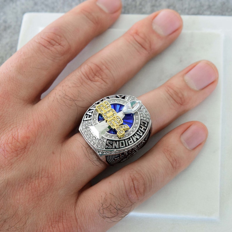 May include: A silver championship ring with a blue center and gold lettering. The ring is encrusted with clear gemstones and features the word "CHAMPIONS" around the band. The ring is on a finger.