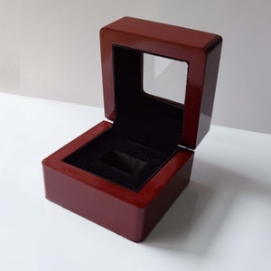 Op de afbeelding: Een donkerrode houten ringendoos met een transparant plastic venster aan de bovenkant. De doos is open en de binnenkant is bekleed met zwart fluweel.