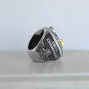 May include: Silver-toned championship ring with the text "2025 CHAMPION" and a football design. The ring features a gold accent and is encrusted with small, clear stones. The ring is designed for a sports championship.