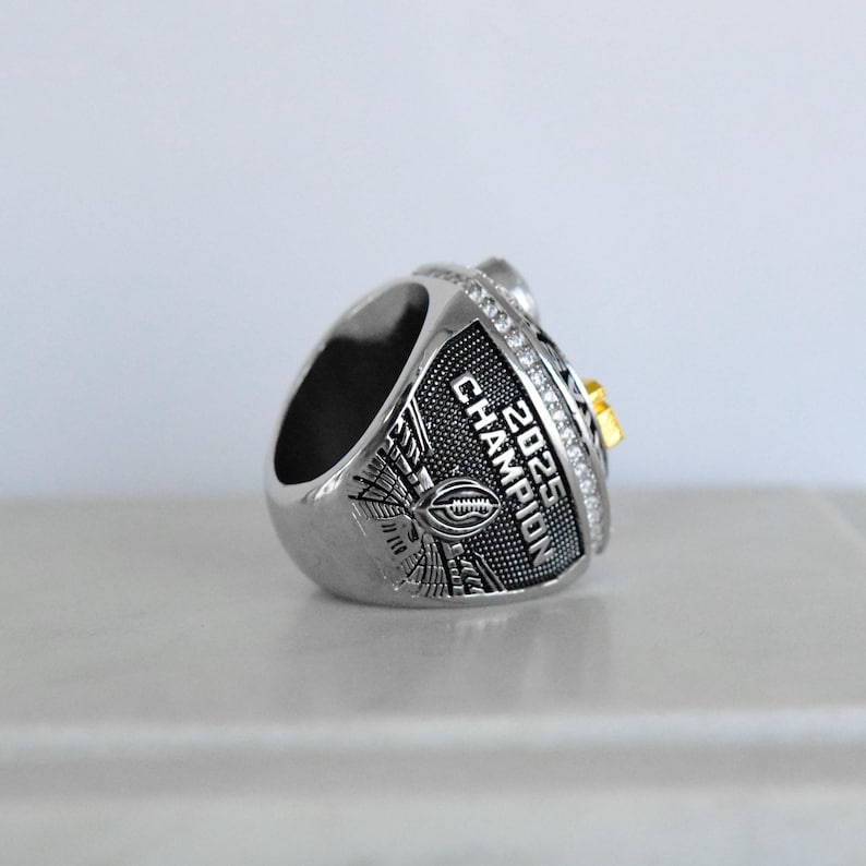 May include: Silver-toned championship ring with the text "2025 CHAMPION" and a football design. The ring features a gold accent and is encrusted with small, clear stones. The ring is designed for a sports championship.