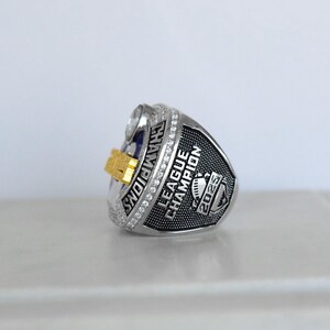 May include: A silver-toned championship ring with the word "CHAMPIONS" engraved on the side. The ring features a gold-colored accent and the words "LEAGUE CHAMPION 2025". The ring is encrusted with small, clear stones.