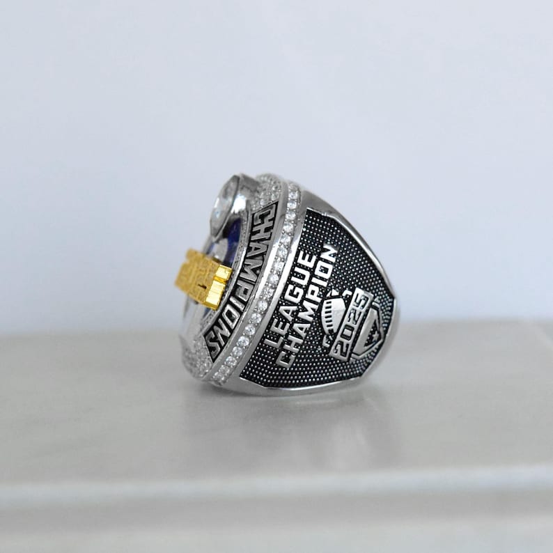 May include: A silver-toned championship ring with the word "CHAMPIONS" engraved on the side. The ring features a gold-colored accent and the words "LEAGUE CHAMPION 2025". The ring is encrusted with small, clear stones.