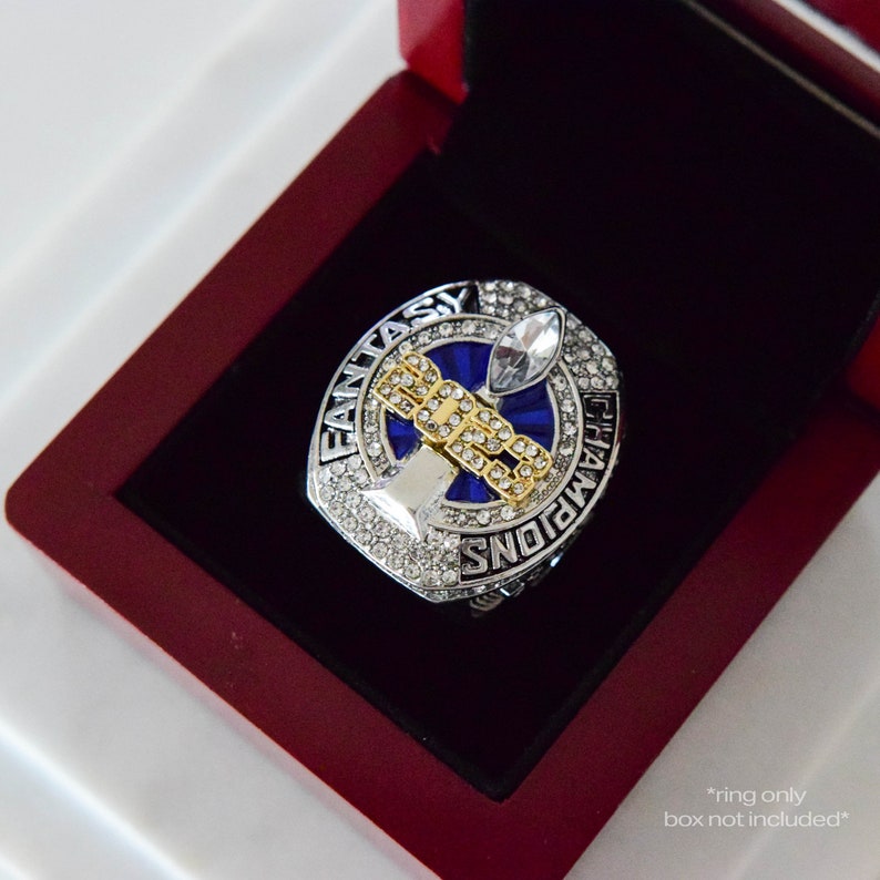 May include: A silver and gold championship ring with blue enamel and rhinestones. The ring features a football trophy design and the words "Fantasy Champions".