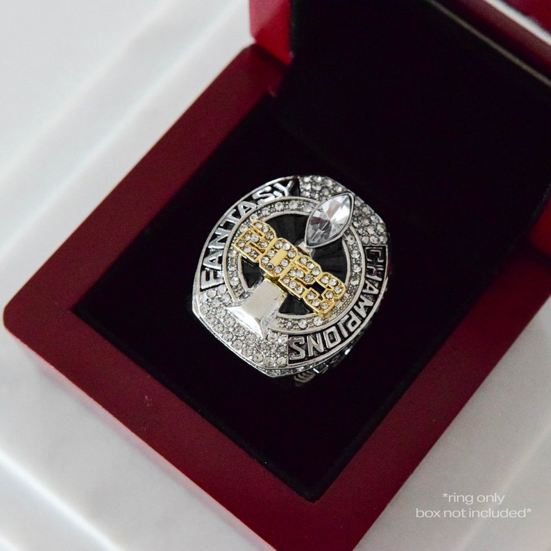 May include: A silver and gold ring with a black background and the words "Fantasy Champions" around the edge. The ring features a silver trophy with a gold base and rhinestones.