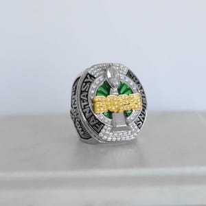 May include: A silver-toned ring with the words "FANTASY CHAMPIONS" engraved around the band. The ring features a green and gold design with the number "2023" in gold lettering. The ring is encrusted with small, clear gemstones.