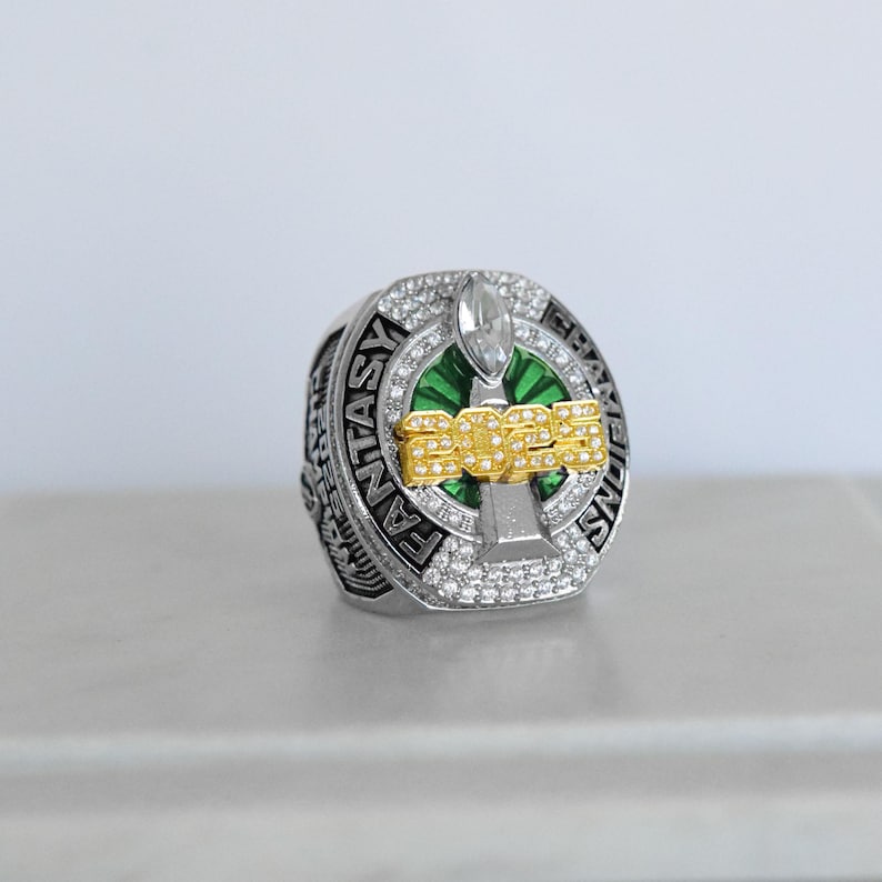 May include: A silver-toned ring with the words "FANTASY CHAMPIONS" engraved around the band. The ring features a green and gold design with the number "2023" in gold lettering. The ring is encrusted with small, clear gemstones.