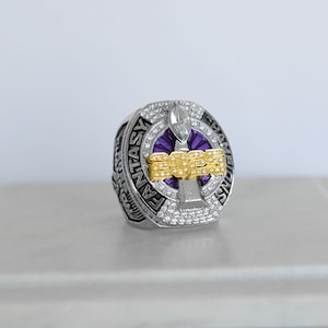 May include: A silver fantasy football championship ring with a purple and gold design. The ring features the year "2023" in gold lettering, surrounded by clear gemstones. The words "FANTASY CHAMPIONS" are engraved on the ring's band.