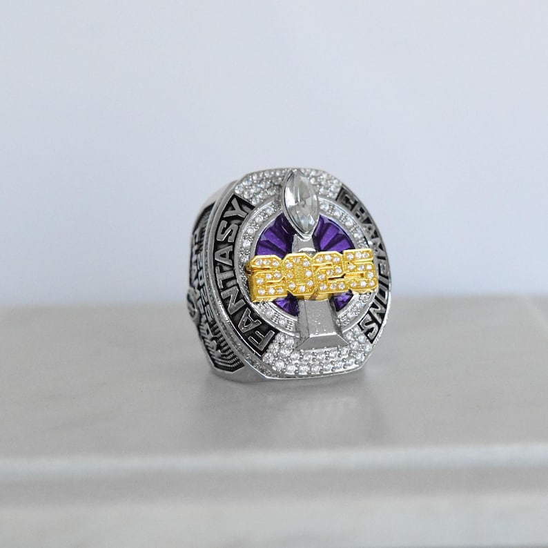 May include: A silver fantasy football championship ring with a purple and gold design. The ring features the year "2023" in gold lettering, surrounded by clear gemstones. The words "FANTASY CHAMPIONS" are engraved on the ring's band.