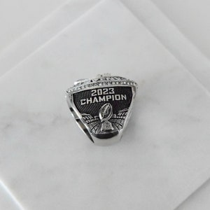 May include: A silver ring with black enamel and rhinestones. The ring features the text "2023 CHAMPION" and a football.
