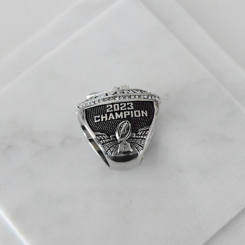 May include: A silver ring with black enamel and rhinestones. The ring features the text "2023 CHAMPION" and a football.